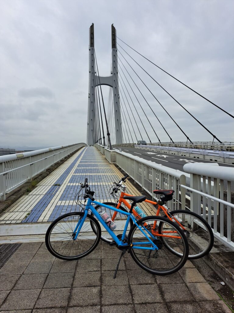 Two bikes, blue and orange parked at the start of a large cabled bridge in Osaka.