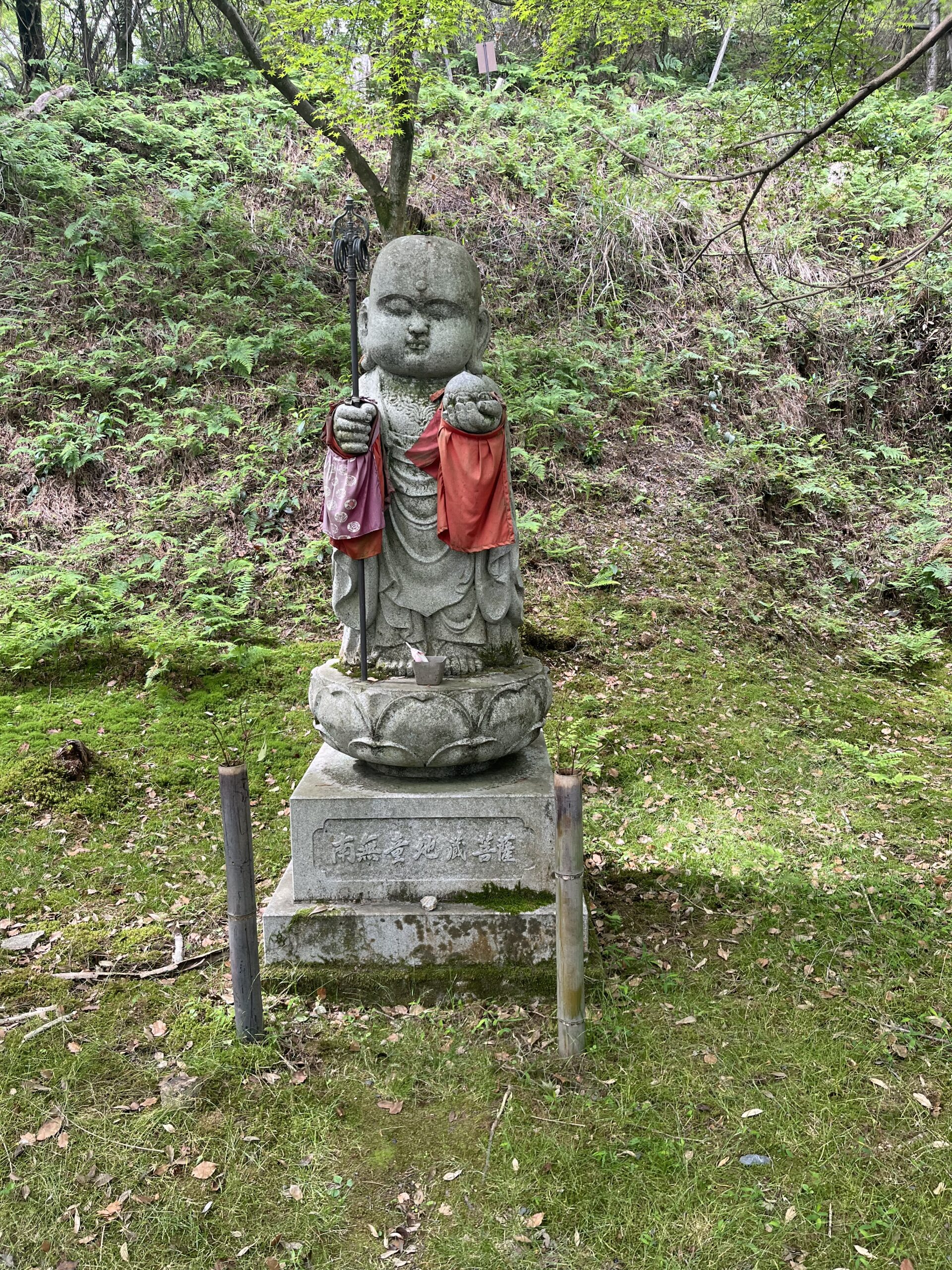 A small stone Jizo statue in the forest, with a red cloth on it's shoulders