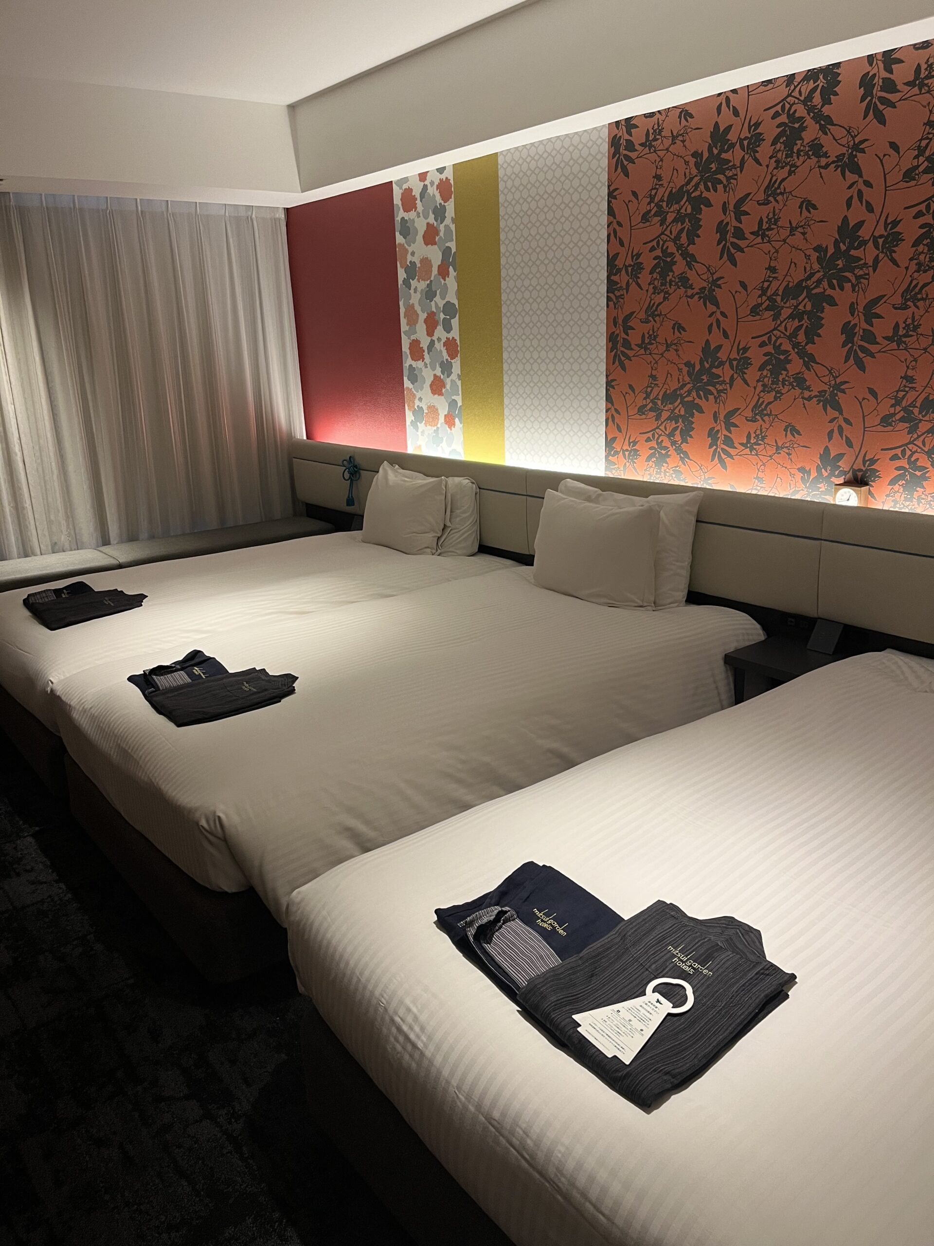 Hotel with three beds and pajamas on the bed with bright colored wall paper on the wall.