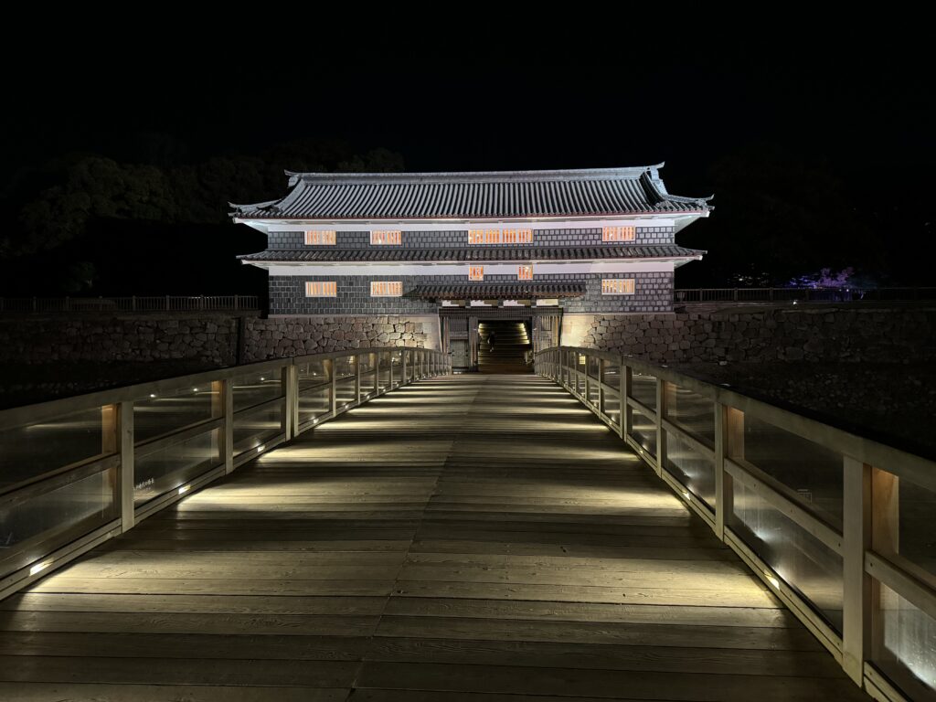 Kanazawa castle bridge entrance at night. Bridge is lit up with gate in background and dark black sky beyond.